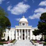 The Alabama State Capitol building in Montgomery with white columns and a gold dome beneath a bright blue sky.