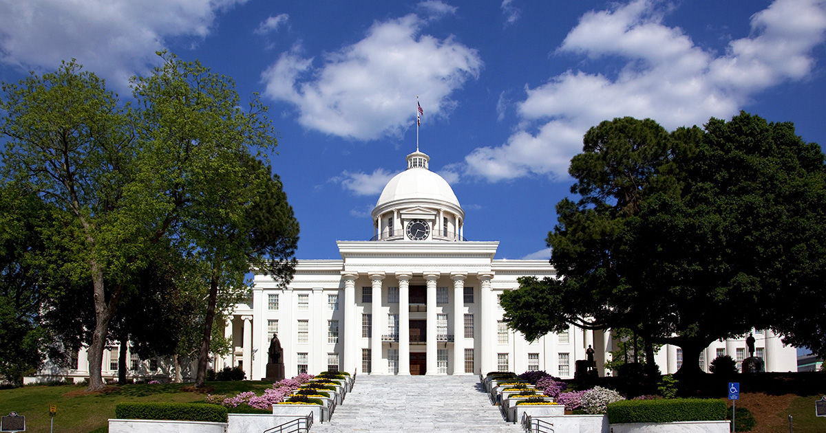 The Alabama State Capitol building in Montgomery with white columns and a gold dome beneath a bright blue sky.