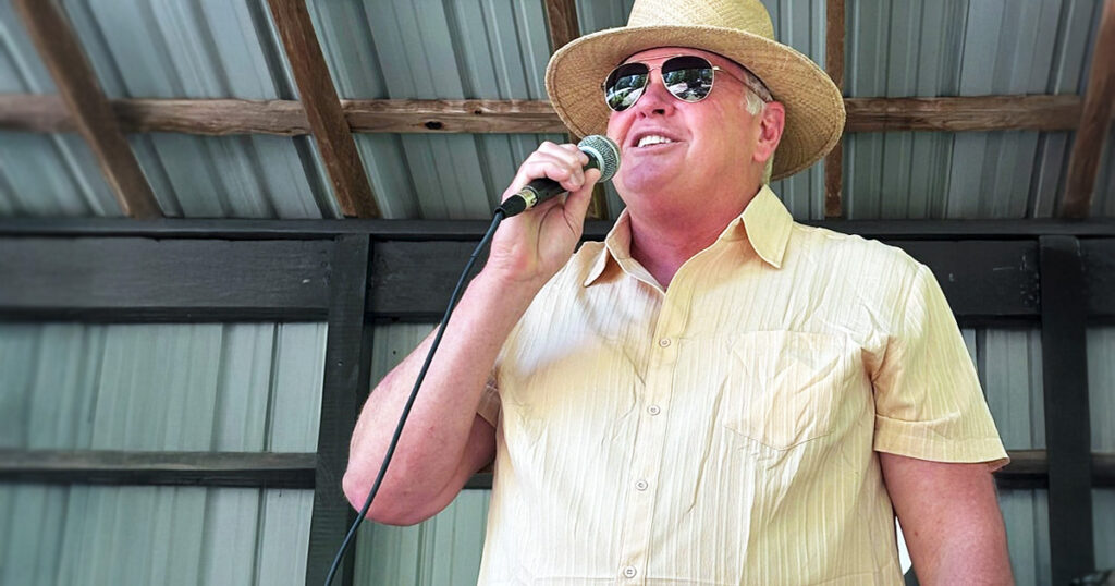 Chad “Chig” Martin speaks into a microphone at a campaign event, wearing a straw hat and sunglasses under a covered outdoor stage.