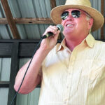 Chad “Chig” Martin speaks into a microphone at a campaign event, wearing a straw hat and sunglasses under a covered outdoor stage.