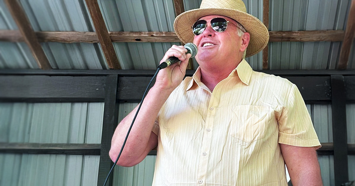 Chad “Chig” Martin speaks into a microphone at a campaign event, wearing a straw hat and sunglasses under a covered outdoor stage.