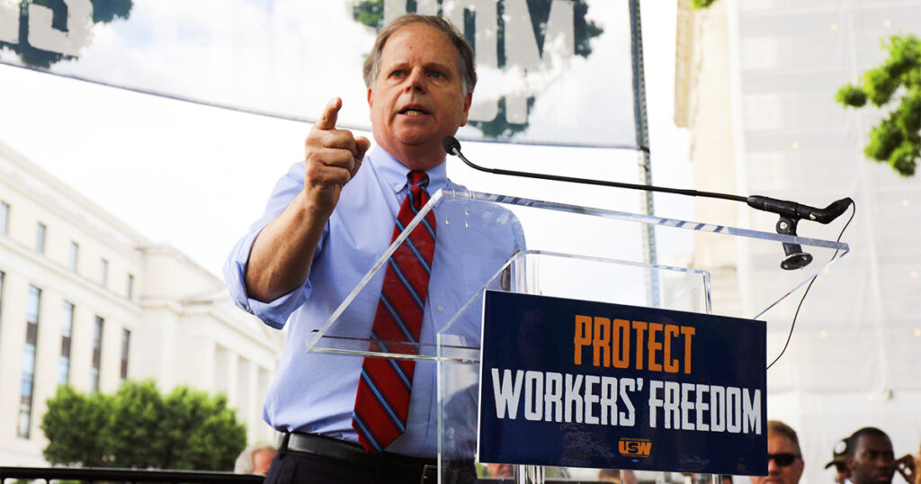 Doug Jones speaks at a rally behind a podium with a “Protect Workers’ Freedom” sign, addressing a crowd outdoors.