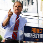 Doug Jones speaks at a rally behind a podium with a “Protect Workers’ Freedom” sign, addressing a crowd outdoors.